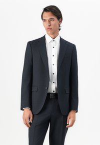 Man wearing dark tailored suit with white dress shirt and black belt, standing against plain white background.