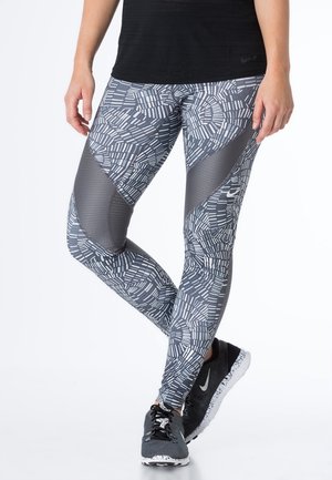 Osoba ubrana w czarno-białe legginsy w wzory i czarne buty sportowe Nike, stojąca z nogami skrzyżowanymi na białym tle.