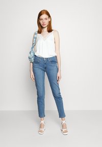 Cămașă de tip camisole cu dantelă albă, blugi skinny de culoare albastru deschis și sandale albe cu bretele. Modelul poartă o poșetă de umăr cu un design albastru.