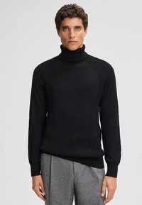 Maglione a collo alto nero a costine con maniche lunghe e un design aderente, realizzato in tessuto testurizzato, abbinato a pantaloni grigi su misura.