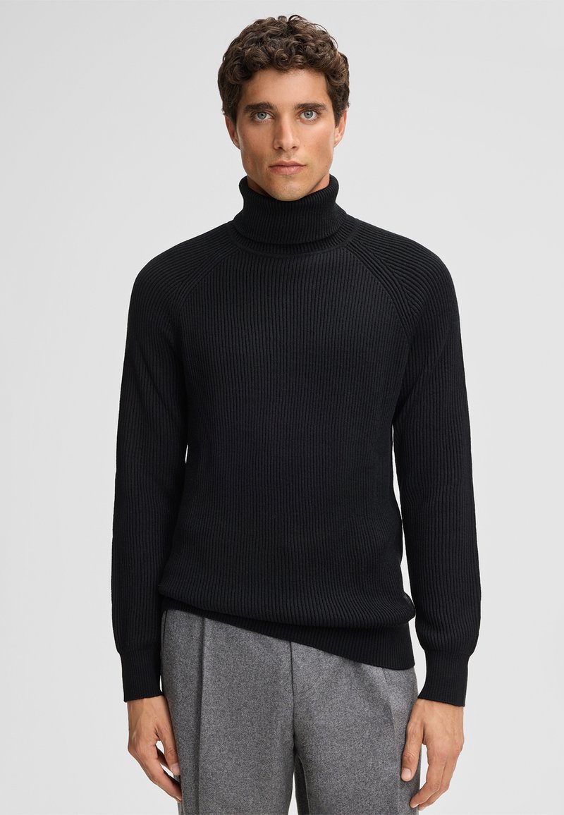 Maglione a collo alto nero a costine con maniche lunghe e un design aderente, realizzato in tessuto testurizzato, abbinato a pantaloni grigi su misura.