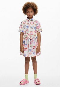 Lyhythihainen mekko, jossa on kuvioitu kangas värikkäine sydämineen ja jälkiruokineen, vyötärö kiristetty ja napit edessä. Vaaleanpunaiset sandaalit ja vihreät sukat.