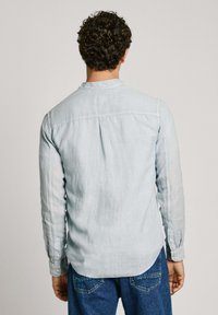 Svetlo modra srajca z dolgimi rokavi, narejena iz mehke tkanine, z ovratnikom na gumb, z volanom na hrbtu in sproščenim krojem. Nosita jo z modrimi kavbojkami.