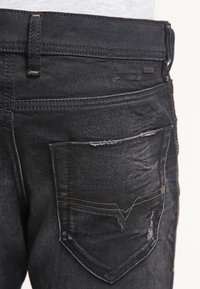Close-up of black denim jeans showing back pocket with mild distressing and visible stitching.