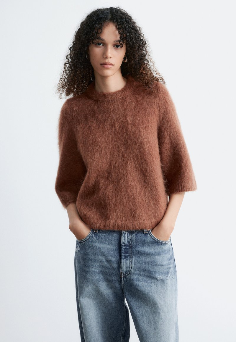Femme aux cheveux bouclés portant un pull marron duveteux à manches trois-quarts et un jean bleu décontracté, mains dans les poches, fond neutre.