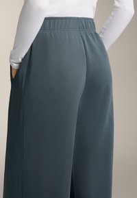 Tyrkysové široké nohavice s rebrovanou textúrou, majú elastický pás a bočné vrecká. Pohľad je zameraný na zadnú časť a bočnú stranu.