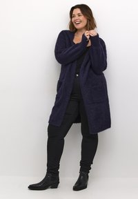 Marineblå fuzzy cardigan med åben front, to lommer, lange ærmer og en afslappet pasform, parret med sorte slim-fit bukser og ankelstøvler.
