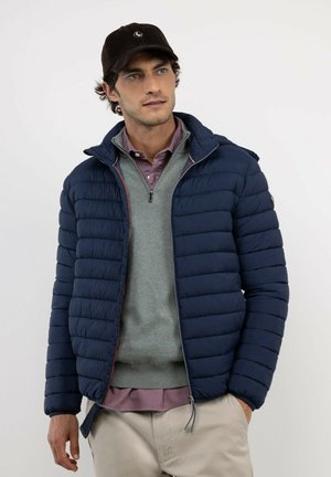 Hombre que lleva una chaqueta azul marino acolchada, un jersey gris con cremallera, camisa morada, pantalones beige y una gorra negra, de pie con las manos en los bolsillos.
