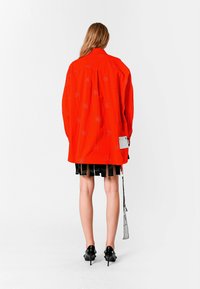 Chaqueta roja de gran tamaño con un patrón bordado, mangas largas, bolsillo lateral y falda plisada negra contrastante. Llevada con zapatos de tacón negros.