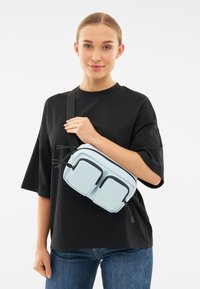 Ljusblå axelremsväska i syntetmaterial med två frampåsar, svarta dragkedjeaccents och justerbar rem.