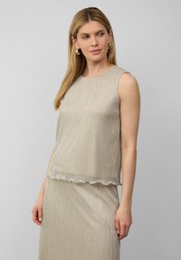 Vrouw met lang blond haar, gekleed in een mouwloze metallic beige top en bijpassende rok, poseert tegen een eenvoudige grijze achtergrond.