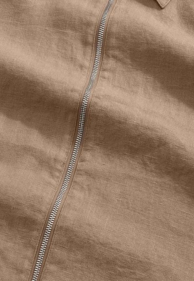 Tissu beige avec une fermeture éclair en métal argenté courant verticalement au centre, montrant une texture détaillée et des coutures.