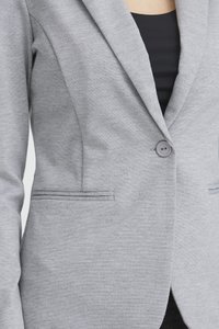 Vrouw draagt een lichtgrijze blazer met een enkele knoop dichtgemaakt over een zwarte top, waarbij het paspelkraagje en de opgestikte zakken van de blazer zichtbaar zijn.