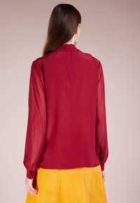 Una donna con lunghi capelli castani indossa una blusa trasparente bordeaux a maniche lunghe e una gonna gialla in suede, vista di spalle.