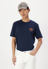 Navy blue T-shirt with short sleeves and a round neck, featuring orange "Wrangler Jeans 1947" text on the chest. Paired with white pants.