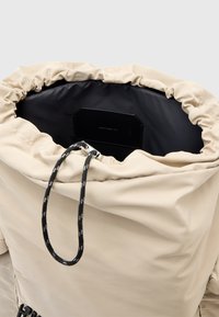 Beige trekzak met een gathered bovenkant, zwarte binnenvoering en een zwart etiket. Gemaakt van een duurzame stof en heeft een stevige trekkoordsluiting.