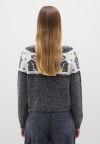 Grau gemusterter Pullover mit rundem Yoke, der einen weißen oberen Bereich mit Tiermotiven und einer flauschigen Textur aufweist. Getragen mit dunkler Hose.