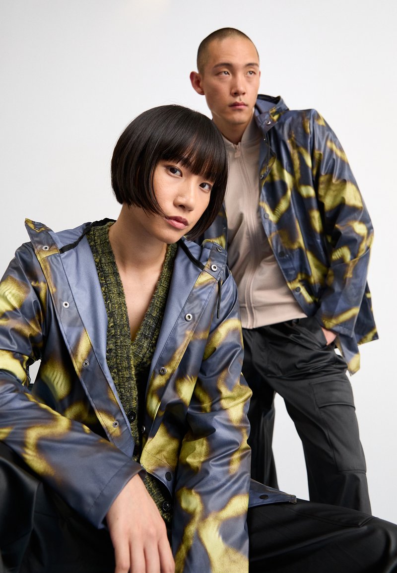 Vestes imperméables bleu et jaune à motifs avec capuches, associées à un pull vert texturé et un pantalon noir. Finition en tissu lisse et brillant.