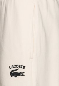 Short beige clair en tissu texturé, avec un logo Lacoste brodé noir et un détail de poche latérale. Ceinture lisse.