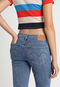 Persona que lleva unos jeans Levi's azules y una camiseta corta con mangas, de corte cropped, con rayas horizontales en rojo, azul, beige y negro, con la mano en el bolsillo trasero.