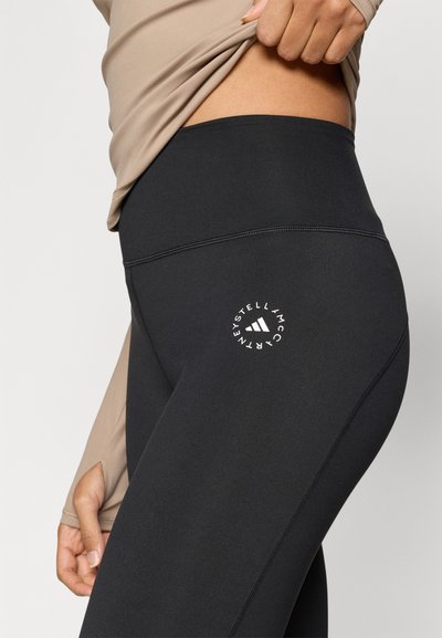 Μαύροι ψηλόμεσοι κολάν με λεία υφή, διακοσμημένοι με λευκό λογότυπο στ ο εξωτερικό μπράτσο και λεπτομέρειες με αντίθετη ραφή.