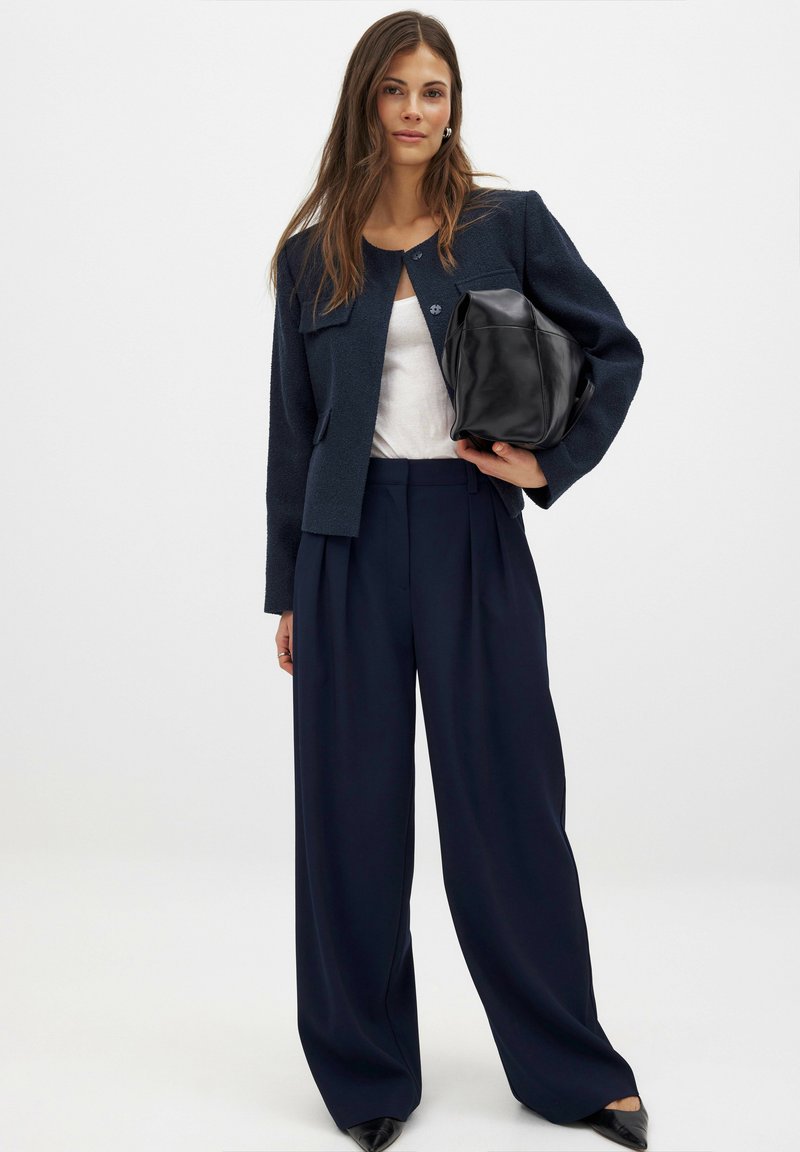 Navy tekstūruotas švarkas su dviem priekinių kišenių, apvalia iškirpte, derinamas su aukšto juosmens, plačiomis kojinėmis jūros mėlynumo kelnių ir juoda odine rankine.