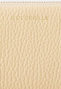 Pouch din piele Coccinelle în crem moale, cu o suprafață texturată, fermoar elegant de culoare aurie și logo embosat pe față.