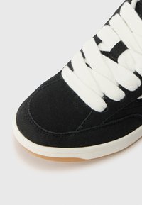 Black suede sneaker with white laces, featuring a rounded toe, stitched accents, and a rubber sole with a brown traction pattern.