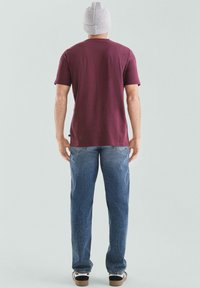 Mężczyzna w szarej dzianinowej czapce, bordowej koszulce, niebieskich dżinsach i czarno-białych trampkach stoi tyłem do kamery.