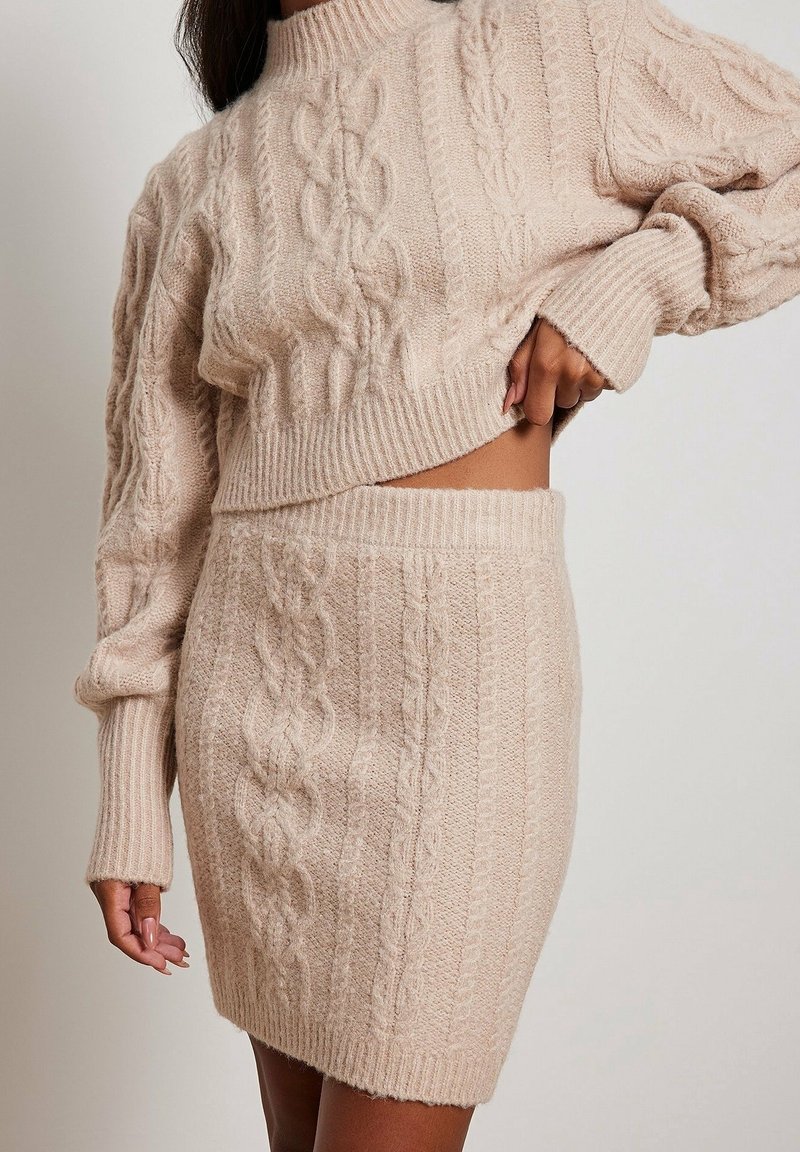 Ensemble pull en maille torsadée beige et jupe. Le pull présente un col montant et une coupe raccourcie ; la jupe adopte une forme ajustée avec des motifs similaires.