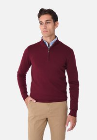 Maglione a maglia bordeaux con colletto rialzato e zip a un quarto. Presenta polsini e orlo a coste, abbinato a pantaloni beige. Texture liscia.