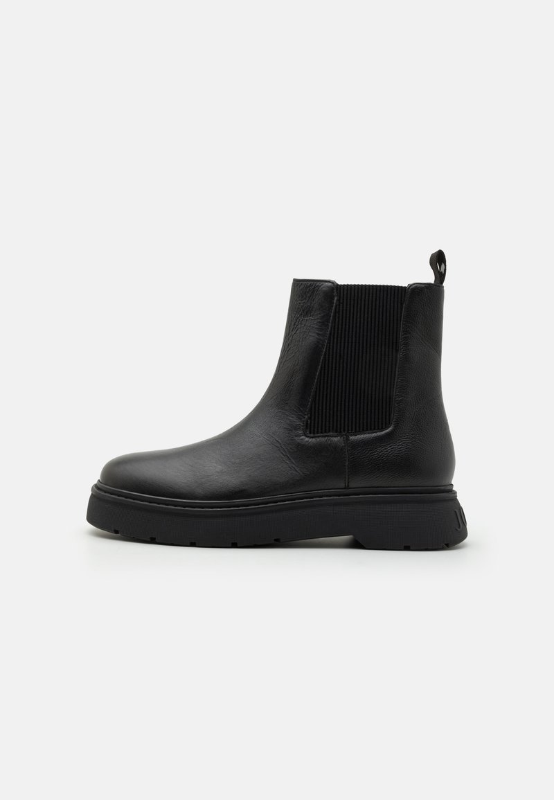 Bottines en cuir noir avec un panneau latéral élastique côtelé, bout rond et semelle épaisse noire. Comprend une languette à l'arrière pour faciliter l'enfilage.