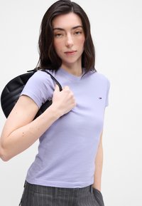 Lyse lilla bomulds t-shirt med rund halsudskæring, korte ærmer og et lille logo på brystet. Kombinationen med en sort skuldertaske og stribede bukser.