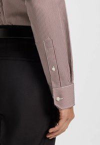 Manche d'une chemise à rayures verticales bordeaux et blanche sur un bras, à côté d'un pantalon noir avec une ceinture visible.