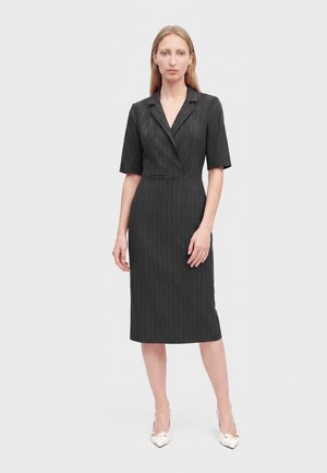 Femme debout portant une robe noire à fines rayures, longueur genou, manches courtes et col cranté, associée à des talons blancs pointus.