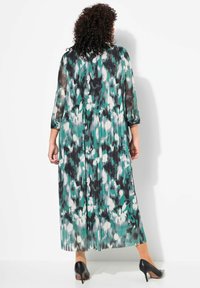 Robe longue plissée à motifs floraux verts, noirs et blancs. Manches translucides trois-quarts et silhouette évasée. Portée avec des escarpins noirs.