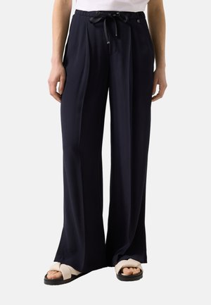 Femme portant un pantalon noir taille haute à jambes larges avec plis à l'avant et taille à cordon, associé à des sandales beige.