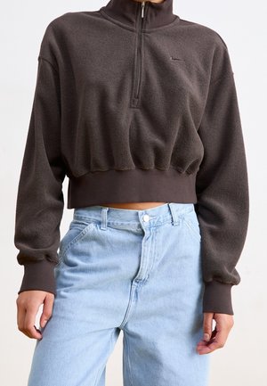 Osoba ubrana w przyciętą, ciemnobrązową bluzę z długim zamkiem do ćwierć długości i długimi rękawami oraz jasnoniebieskie, wysokowalne dżinsy.