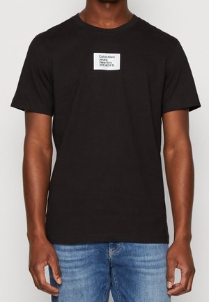 Schwarzes Baumwoll-T-Shirt mit kurzen Ärmeln, ausgestattet mit einem rechteckigen weißen Etikett, auf dem "Calvin Klein Jeans New York 205 W39 St." gedruckt ist.