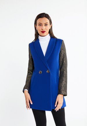 Femeie care poartă un blazer albastru fără mâneci, cu mâneci lungi negre texturate și un guler alb, stând în fața unui fundal alb.