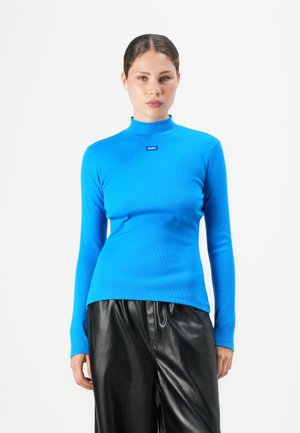 Vrouw in een felblauwe lange mouw top met een hoge hals en een klein "HUGO" logo, met zwarte leren broek, staande tegen een witte achtergrond.