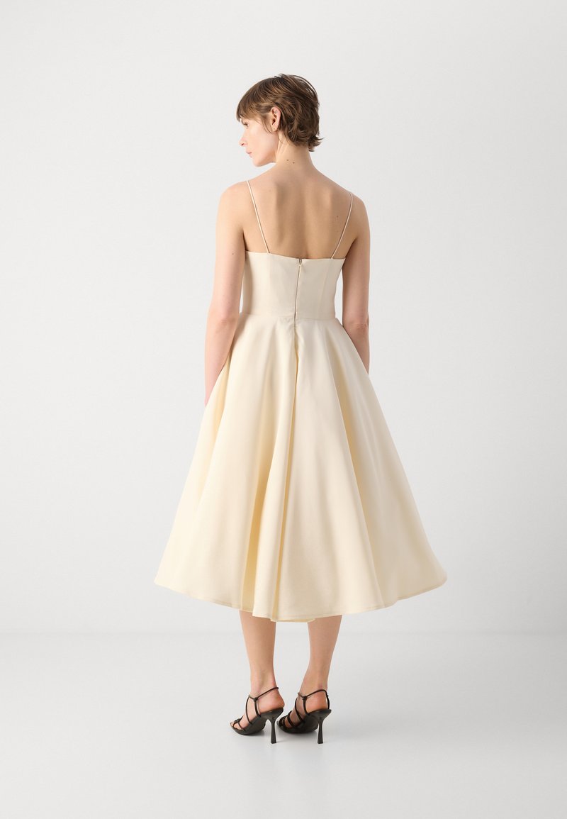 Ein knielanges, cremefarbenes Kleid mit dünnen Trägern, glattem Stoff, ausgestelltem Schnitt und einem verdeckten Reißverschluss hinten, kombiniert mit schwarzen Riemchenabsätzen.