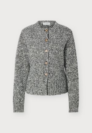 Femme portant un cardigan gris chiné boutonné, un pantalon large noir et des chaussures noires, debout devant un fond clair uni.