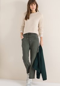 Femme portant un pull beige et un pantalon vert, tenant une veste vert foncé, debout contre un mur beige clair.