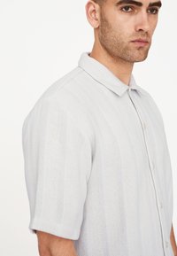 Polo de manga corta gris claro, hecho de una tela de punto texturizada con rayas horizontales y cierre de botones en la parte delantera. Diseño clásico de cuello.