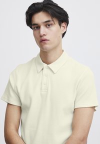 Polo-skjorte i lys beige, laget av teksturert stoff. Har krage, korte ermer og tre frontknapper. Enkel, strømlinjeformet design.