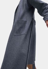 Cappotto lungo grigio strutturato con maniche lunghe, tasche laterali e vita legata. Il tessuto presenta un sottile motivo a spina di pesce.