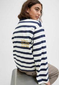 A person with wavy brown hair wears a striped shirt displaying a message on the back. They pose calmly on a metal surface.