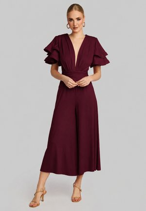 Donna bionda che indossa una tuta bordeaux scuro con maniche corte arricciate e gambe larghe, in piedi con le mani giunte, tacchi beige.