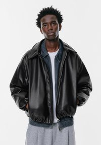 Črna umetna usnjena bomber jakna z visokim ovratnikom in elastičnimi manšetami. Ima zadrgo spredaj in plastev denima na dnu.